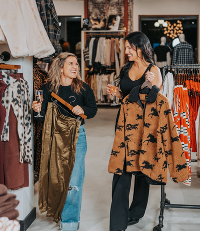 Two women in a clothing store holding drinks and a patterned coat.
